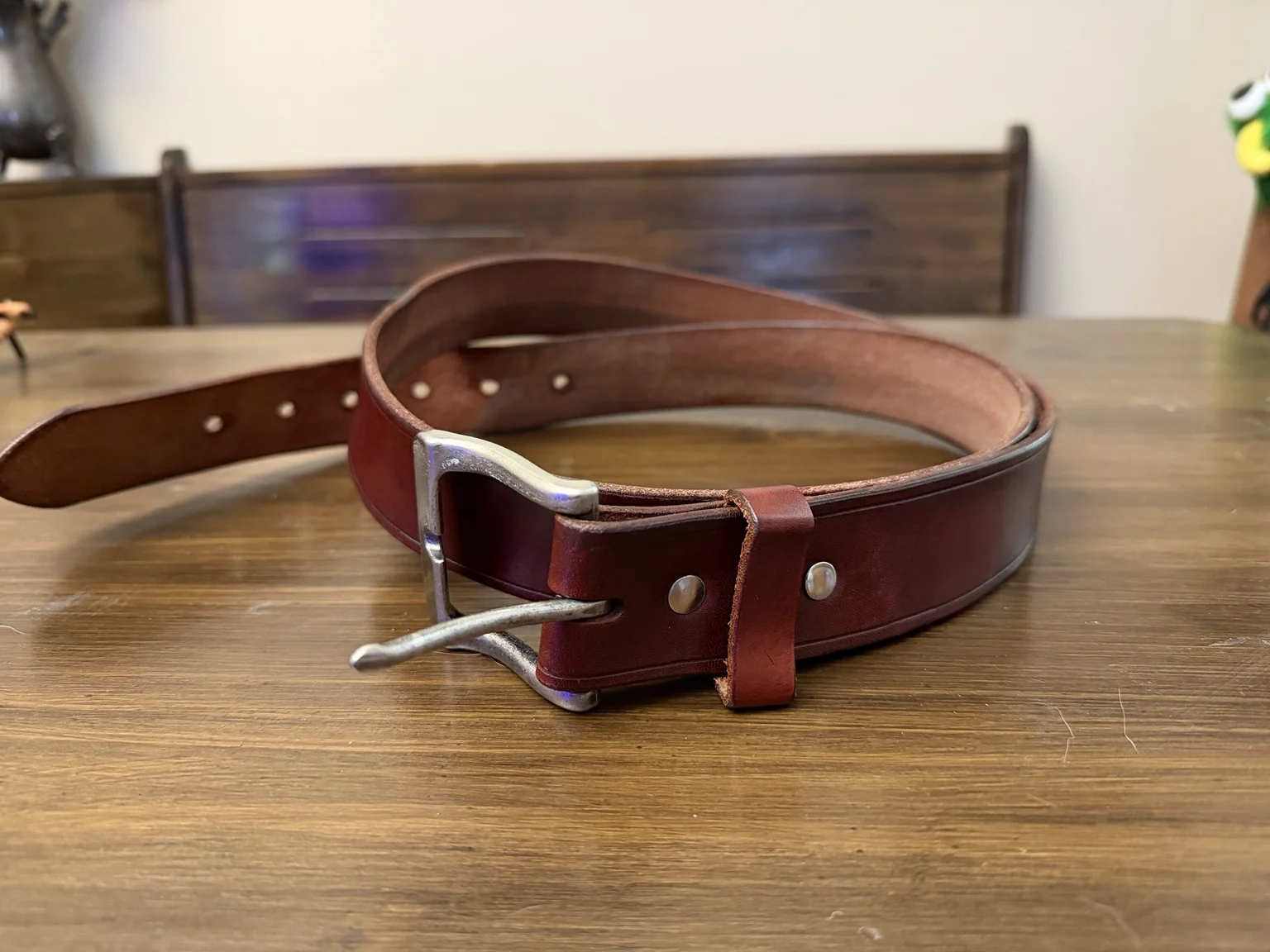 The reddish-brown leather belt Alex made, loosely coiled on a wooden table with its silver-colored buckle at the front.